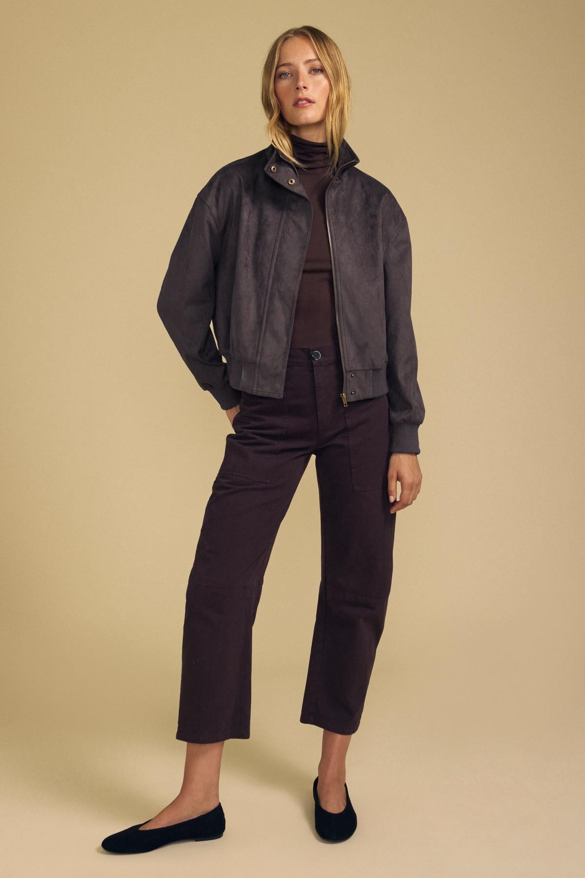 A woman stands against a beige background wearing the QUINCY FAUX SUEDE BOMBER JACKET by Velvet by Graham & Spencer layered over a brown turtleneck, paired with cropped brown pants and black flats for a modern silhouette.-42157575536833