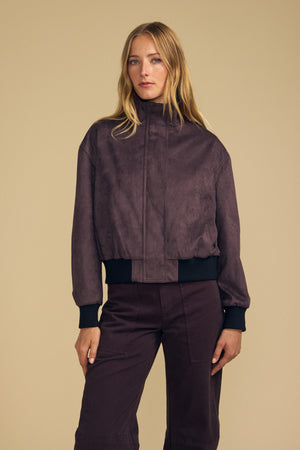 A woman stands against a beige background wearing the QUINCY FAUX SUEDE BOMBER JACKET by Velvet by Graham & Spencer and matching high-waisted pants with a modern silhouette, her left hand resting at her side.