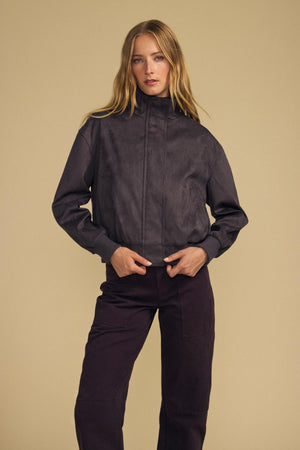 A woman stands against a beige background wearing the QUINCY FAUX SUEDE BOMBER JACKET and matching pants from Velvet by Graham & Spencer, her hands resting near the front of the jacket. The faux suede fabric and modern silhouette lend a touch of contemporary elegance to her look.