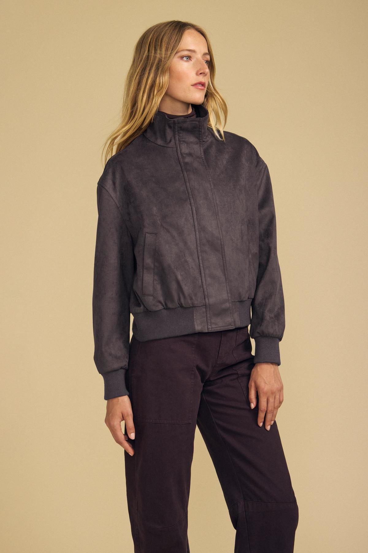 A woman models the QUINCY FAUX SUEDE BOMBER JACKET by Velvet by Graham & Spencer in dark gray, featuring a high collar and matched with coordinating pants, standing against a plain beige background. The modern silhouette of this faux suede ensemble lends contemporary elegance to her look.-42157575438529