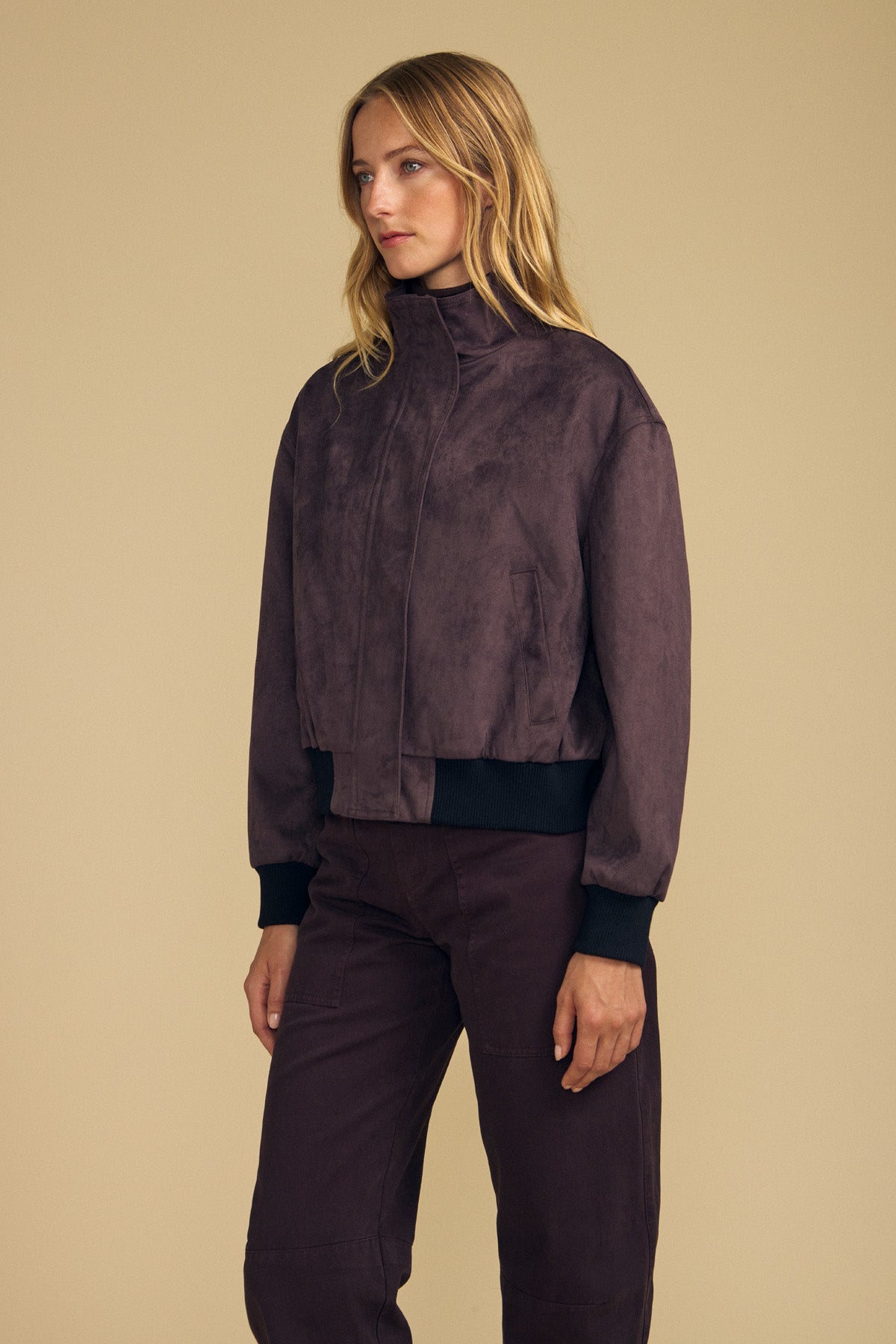 A woman stands against a beige background wearing the QUINCY FAUX SUEDE BOMBER JACKET by Velvet by Graham & Spencer, styled with matching brown pants and looking to the side.