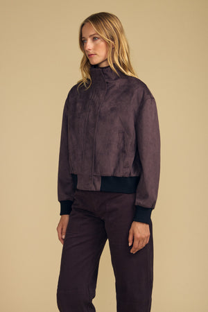 A woman stands against a beige background wearing the QUINCY FAUX SUEDE BOMBER JACKET by Velvet by Graham & Spencer, styled with matching brown pants and looking to the side.