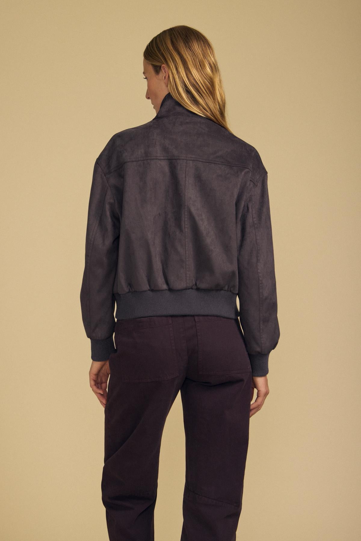 A person with long blond hair is standing with their back to the camera, wearing the QUINCY FAUX SUEDE BOMBER JACKET by Velvet by Graham & Spencer and dark pants against a plain beige background.