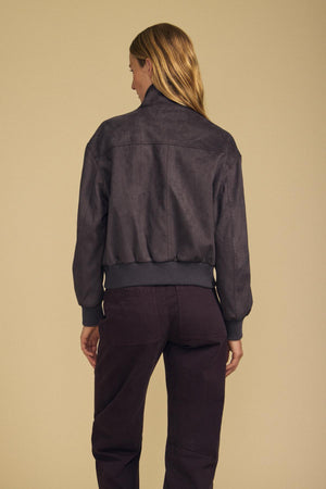 A person with long blond hair is standing with their back to the camera, wearing the QUINCY FAUX SUEDE BOMBER JACKET by Velvet by Graham & Spencer and dark pants against a plain beige background.