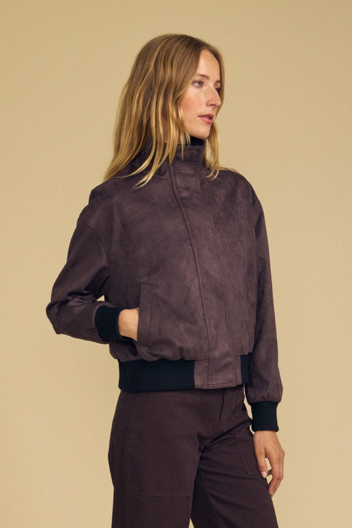 A woman stands against a beige background with her hands in her pockets, wearing the QUINCY FAUX SUEDE BOMBER JACKET by Velvet by Graham & Spencer—an elegant dark brown jacket featuring black cuffs—paired with brown pants, as she looks off to the side.-41964710854849