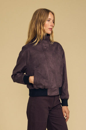 A woman stands against a beige background with her hands in her pockets, wearing the QUINCY FAUX SUEDE BOMBER JACKET by Velvet by Graham & Spencer—an elegant dark brown jacket featuring black cuffs—paired with brown pants, as she looks off to the side.