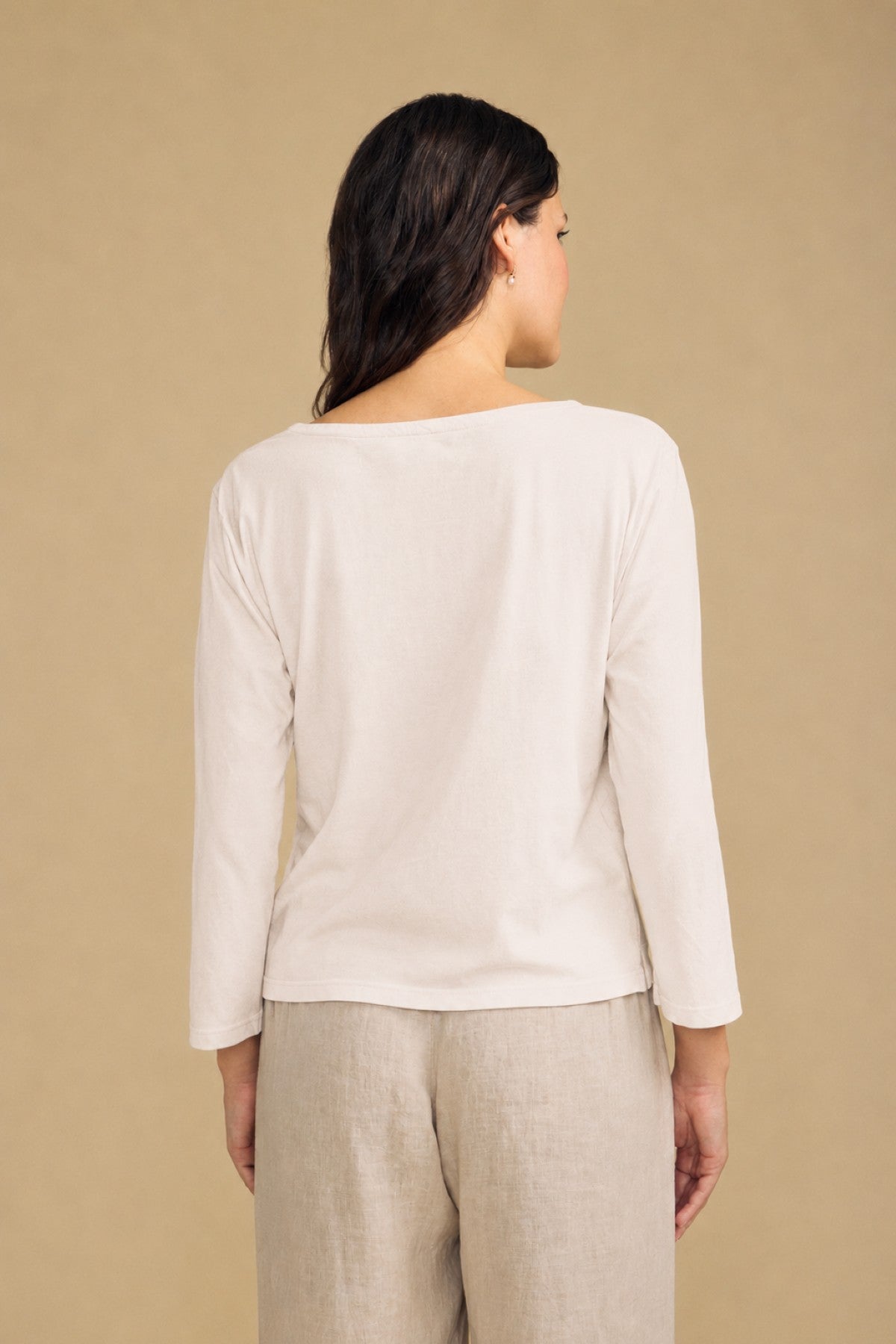 A person stands facing away, wearing the Velvet by Graham & Spencer GABI TEE—a long-sleeved, off-white cotton jersey top with a boat neckline—paired with light beige pants against a plain beige background.