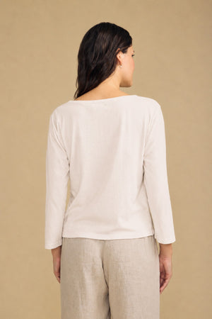 A person stands facing away, wearing the Velvet by Graham & Spencer GABI TEE—a long-sleeved, off-white cotton jersey top with a boat neckline—paired with light beige pants against a plain beige background.
