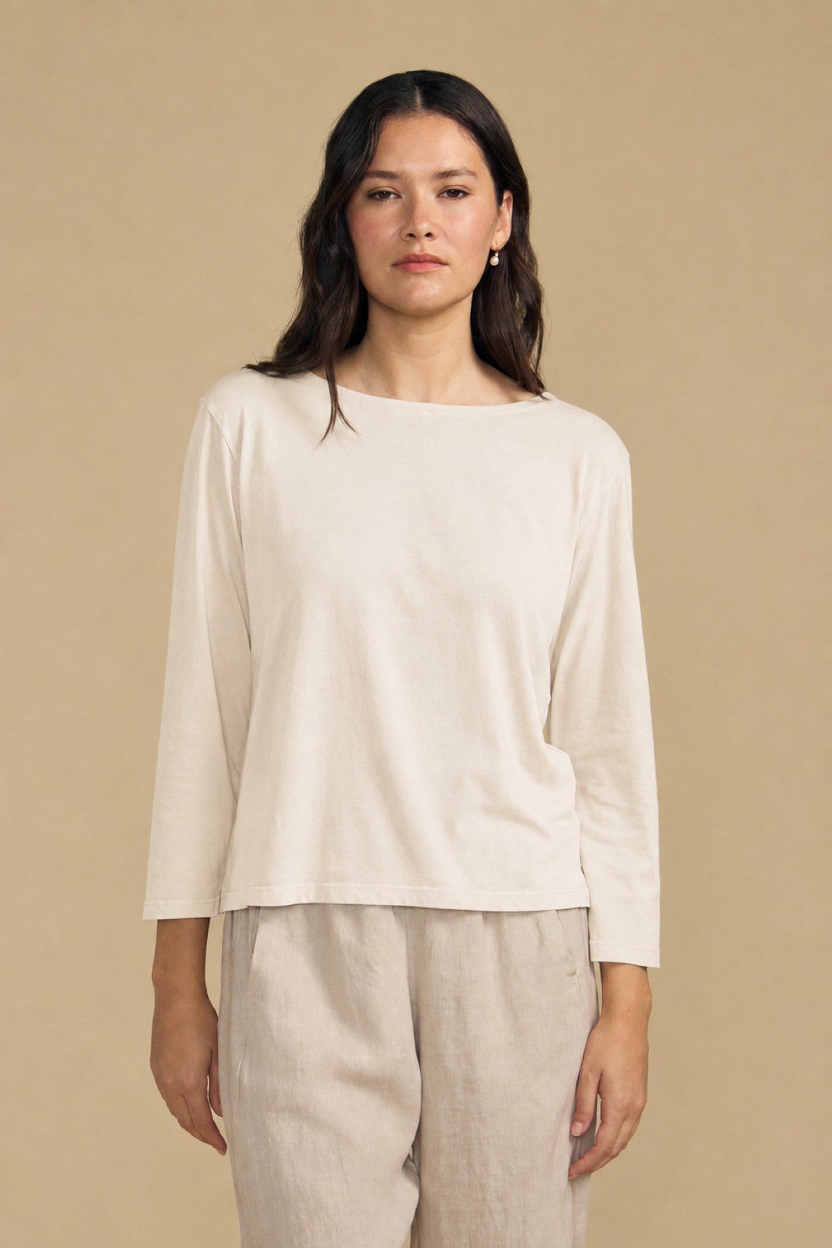 A person with long dark hair wears the Velvet by Graham & Spencer GABI TEE, a loose, light beige cotton jersey top with a boat neckline, paired with matching pants and standing against a plain beige background.