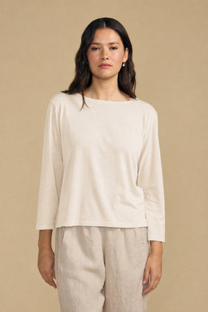 A person with long dark hair wears the Velvet by Graham & Spencer GABI TEE, a loose, light beige cotton jersey top with a boat neckline, paired with matching pants and standing against a plain beige background.