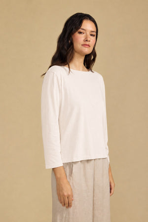 A woman with long, dark hair wears the GABI TEE by Velvet by Graham & Spencer—a loose, light beige cotton jersey top with a boat neckline—paired with matching cropped, wide-leg pants as she stands against a plain beige background.