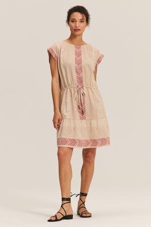 A woman models the CHANTEL DRESS by Velvet by Graham & Spencer, a woven cotton jacquard piece with short sleeves, red geometric patterns, a drawstring waist, and black lace-up sandals, set against a plain background.