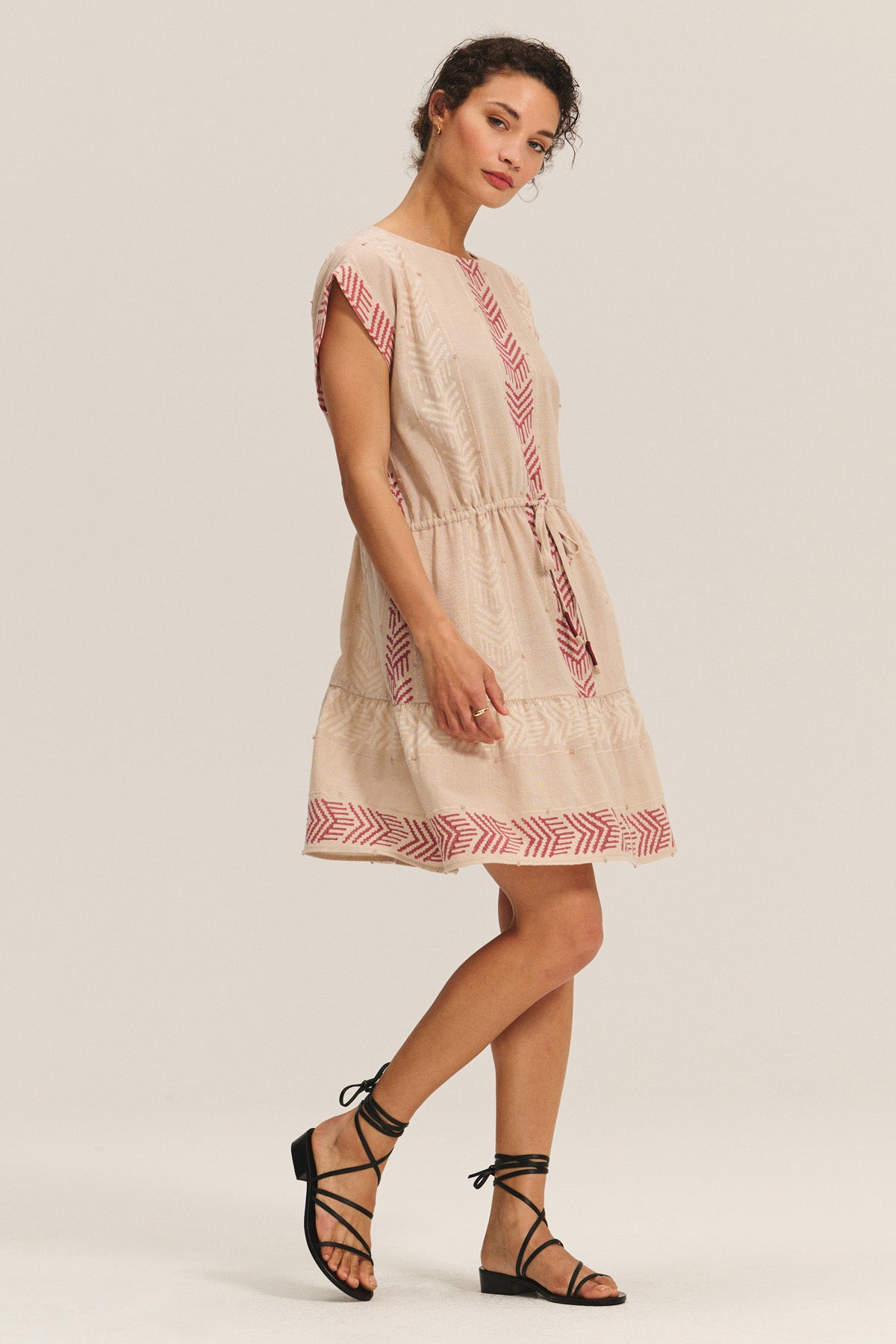 A woman wears the CHANTEL DRESS by Velvet by Graham & Spencer, a short-sleeve woven cotton jacquard dress with red geometric patterns and a drawstring waist, paired with black lace-up sandals.