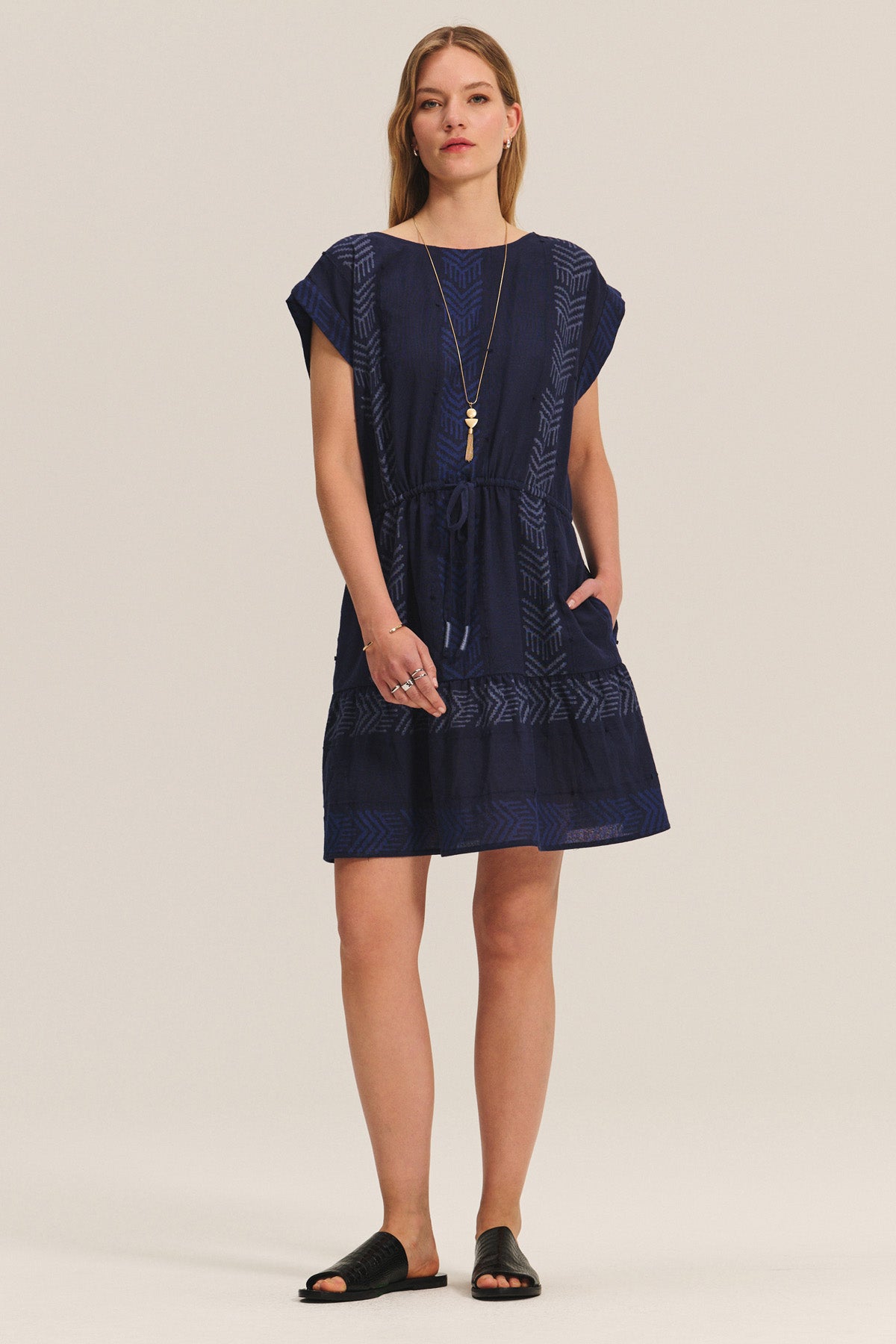 A woman stands facing forward wearing the CHANTEL DRESS by Velvet by Graham & Spencer, a navy short-sleeve woven cotton jacquard dress with geometric patterns, a drawstring waist, and pockets. She pairs it with a long necklace and black sandals.-40732758114497
