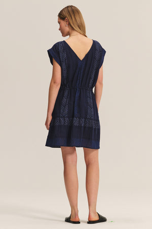 A woman faces away, wearing the CHANTEL DRESS by Velvet by Graham & Spencer—a navy blue short-sleeve woven cotton jacquard dress with a V-back, geometric patterns, and black slip-on shoes.