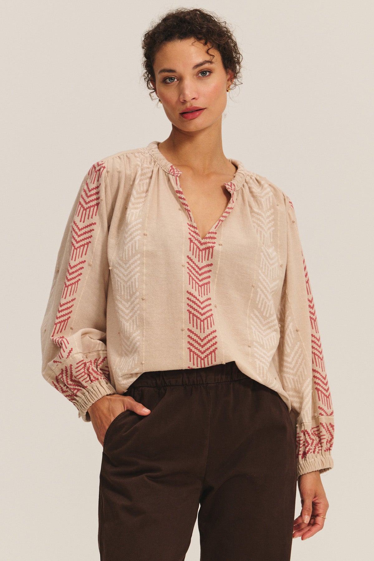 A woman wears the EFFIE TOP by Velvet by Graham & Spencer—a flowy cotton jacquard blouse with red geometric patterns and long sleeves—paired with dark brown pants, standing against a plain background.