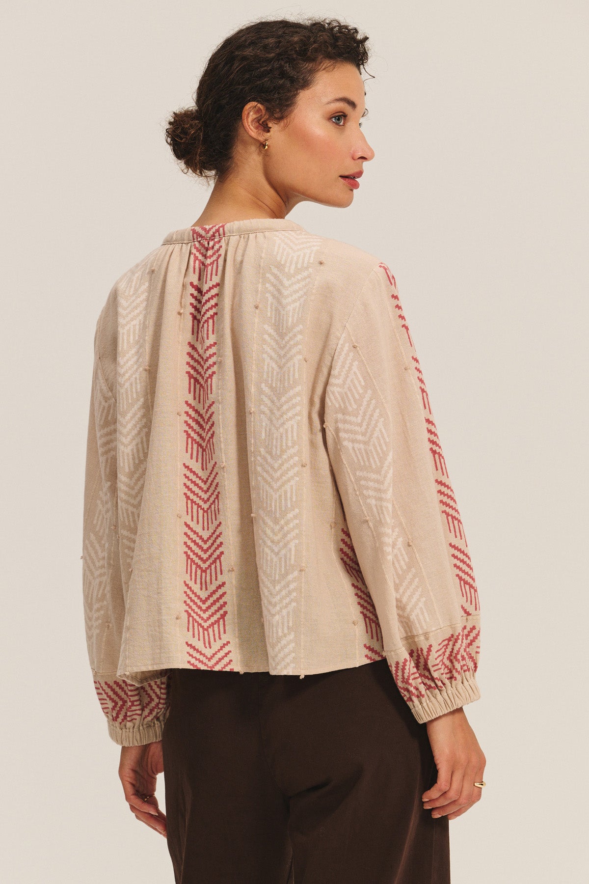 A woman is seen from the back wearing the Velvet by Graham & Spencer EFFIE TOP, a beige blouse with red and white geometric patterns, paired with dark pants that accentuate her feminine silhouette.-40732809986241