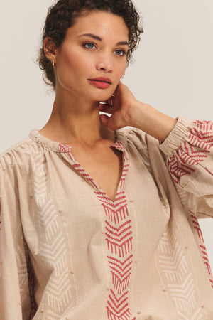 A woman with short curly hair wears the EFFIE TOP by Velvet by Graham & Spencer— a cotton jacquard blouse with red geometric embroidery—standing against a neutral background, one hand touching her neck and highlighting her feminine silhouette.