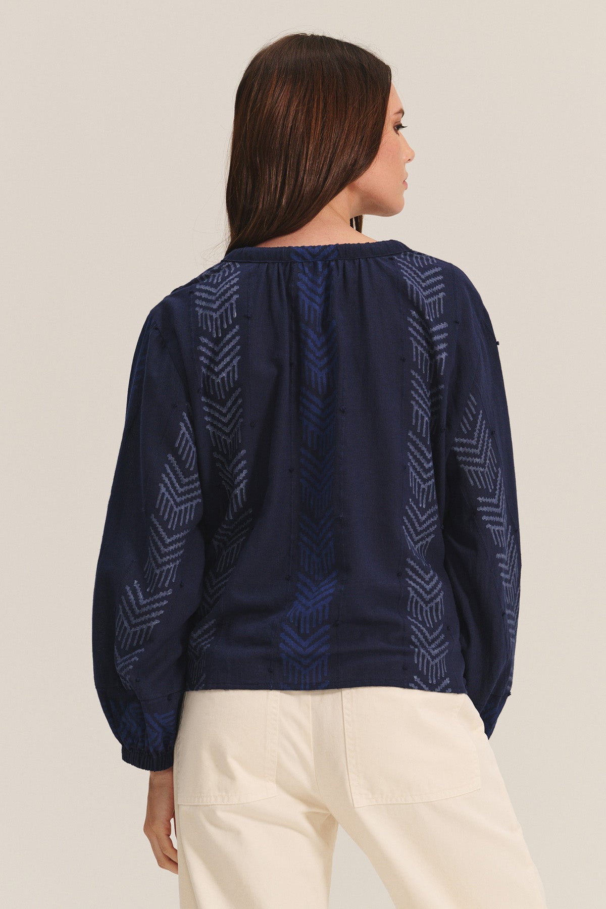 A woman faces away, wearing the EFFIE TOP by Velvet by Graham & Spencer—a navy cotton jacquard blouse with geometric patterns—paired with off-white pants, showcasing a feminine silhouette against a plain light background.