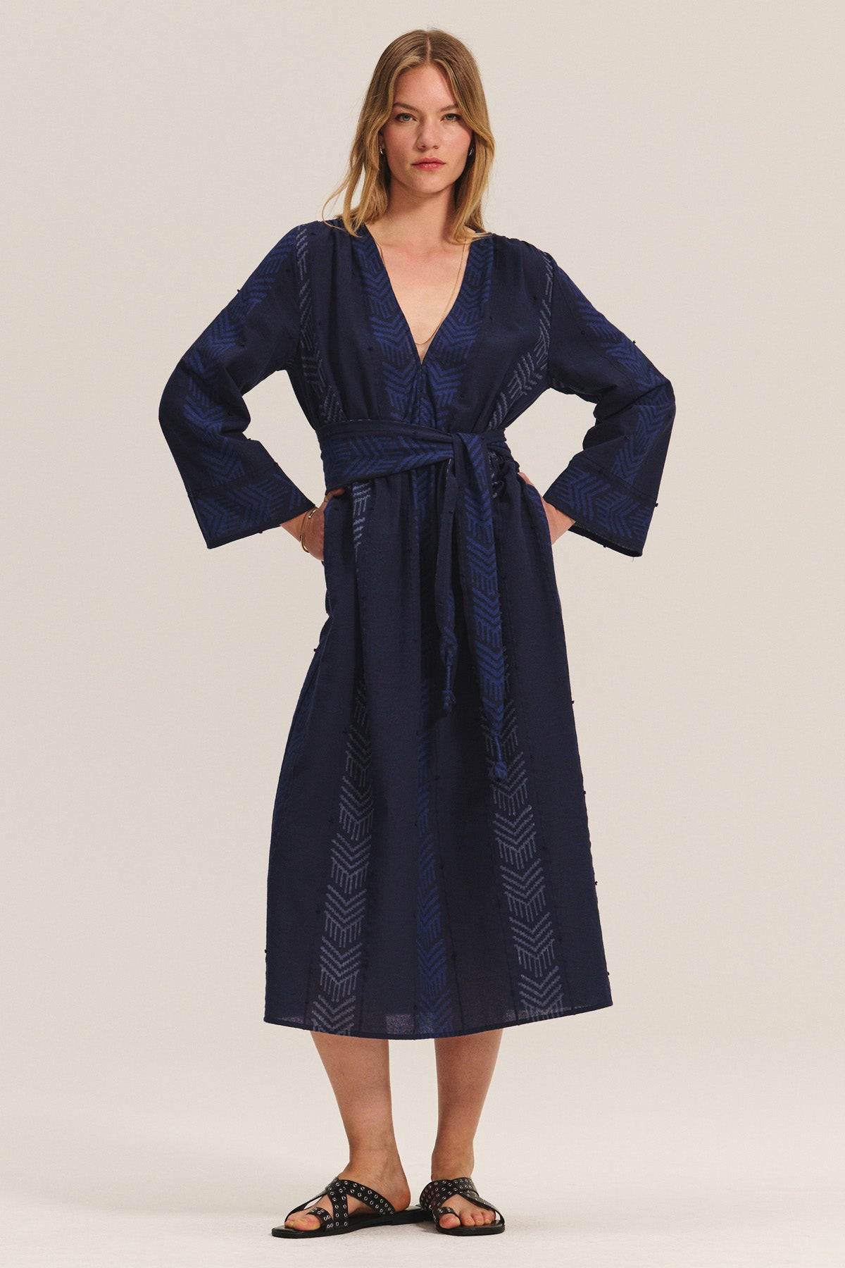 A woman poses with hands on hips, wearing the ELDORA DRESS by Velvet by Graham & Spencer—a navy blue, long-sleeve woven cotton dress with a V-neckline, geometric patterns, and detachable belt—paired with black studded sandals.-40732507832513