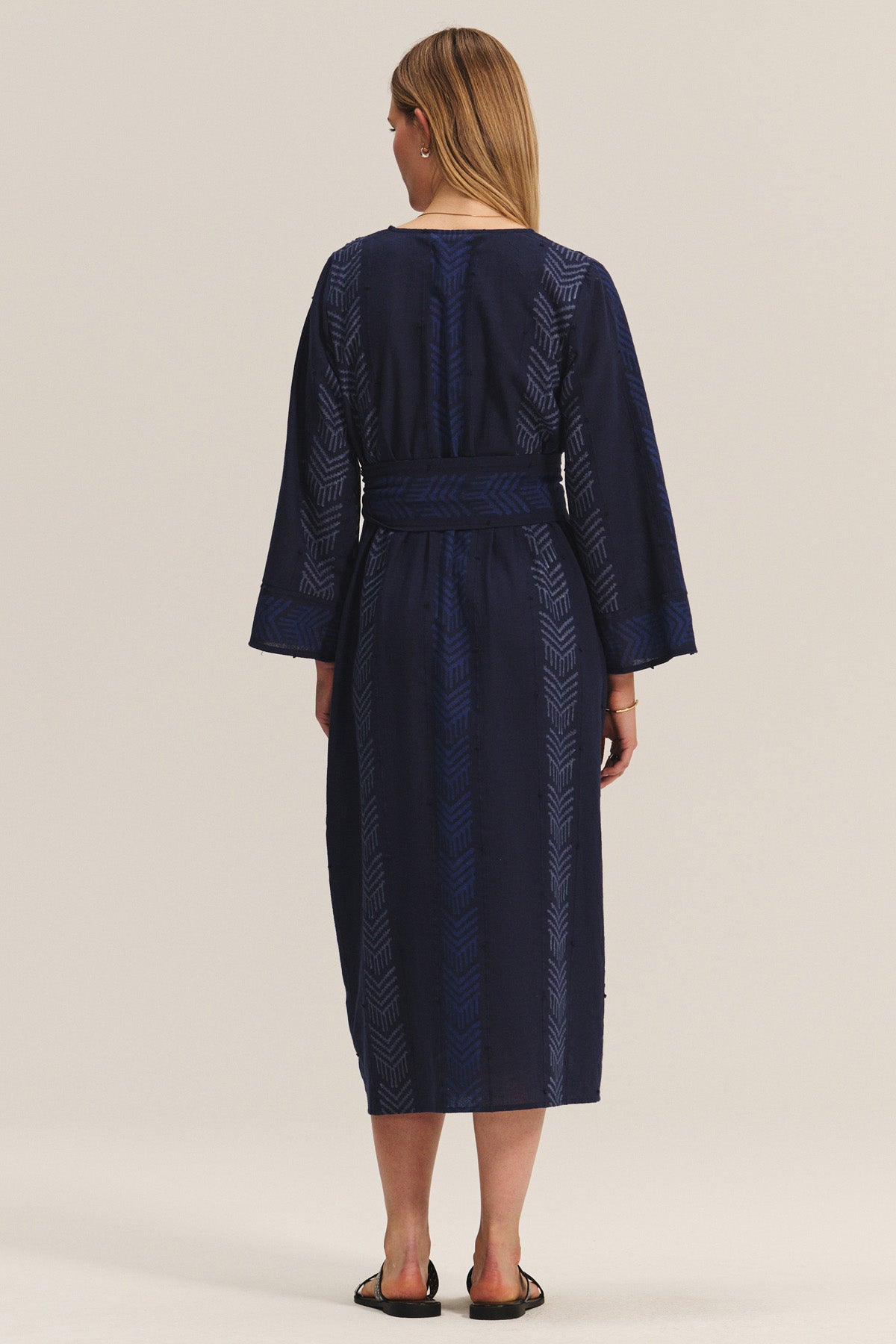 A woman faces away wearing the ELDORA DRESS by Velvet by Graham & Spencer—a navy woven cotton dress with a geometric print, V-neckline, and waist tie—styled with black sandals.-40732507799745