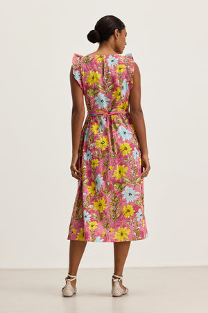 A woman faces away, wearing the ELARIA DRESS by Velvet by Graham & Spencer—a pink floral cotton dress with flutter sleeves, ruffled shoulders, a belt, and side seam pockets—paired with white flat sandals.