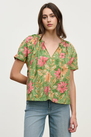 A woman poses against a simple backdrop, wearing the IKRAM TOP: a relaxed fit green floral blouse with pink flowers by Velvet by Graham & Spencer, crafted from lightweight cotton, paired with blue jeans.