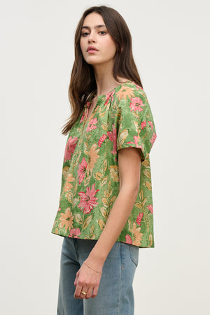 A woman in the IKRAM TOP, a relaxed fit floral green blouse by Velvet by Graham & Spencer, paired with jeans, stands against a plain background, looking slightly to the side.