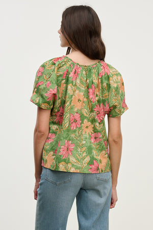 A person stands with their back turned, wearing the Velvet by Graham & Spencer IKRAM TOP—a lightweight cotton blouse featuring a green and pink floral pattern and short sleeves—paired with blue jeans.