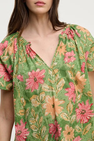 A person wearing the IKRAM TOP by Velvet by Graham & Spencer, a relaxed fit green floral-print top featuring pink and orange flowers on a plain background, made from lightweight cotton.