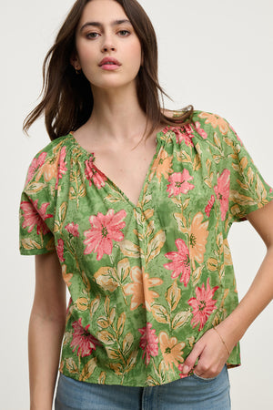 Someone is wearing the IKRAM TOP by Velvet by Graham & Spencer, a relaxed fit green floral cotton blouse with pink and orange flowers and a ruffle neckline. It’s perfectly matched with blue jeans against a plain background.
