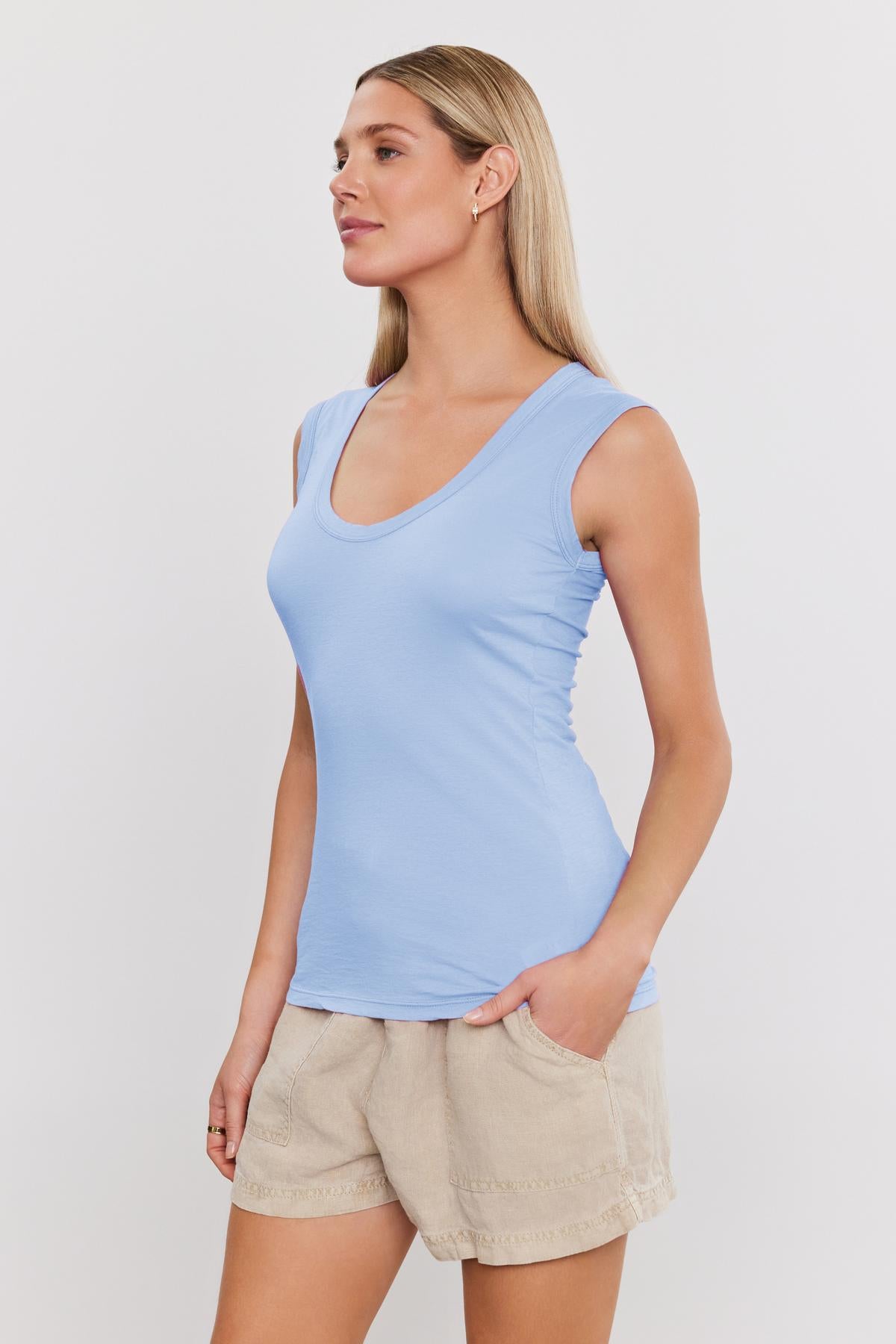   A woman wearing a Velvet by Graham & Spencer Estina Gaucy Whisper fitted tank top in light blue, paired with beige shorts, standing profile to the camera with a neutral expression. 