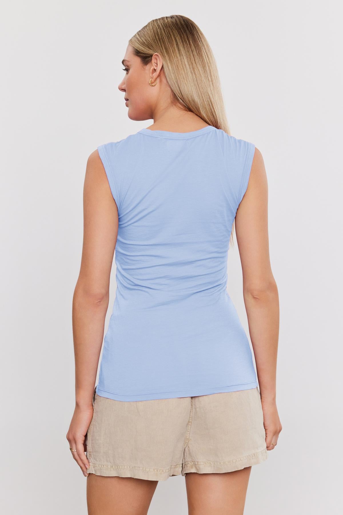   A woman viewed from the back, wearing a blue ESTINA GAUZY WHISPER FITTED TANK TOP and beige shorts, standing against a white background. Brand Name: Velvet by Graham & Spencer 