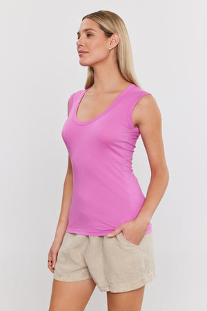 A woman with long blonde hair in the ESTINA TANK TOP by Velvet by Graham & Spencer and beige shorts stands with her hands in her pockets against a plain white background.