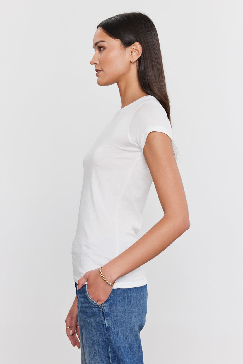 A woman with long, straight hair stands in profile, wearing a JEMMA TEE by Velvet by Graham & Spencer and blue jeans with her hand in her pocket.