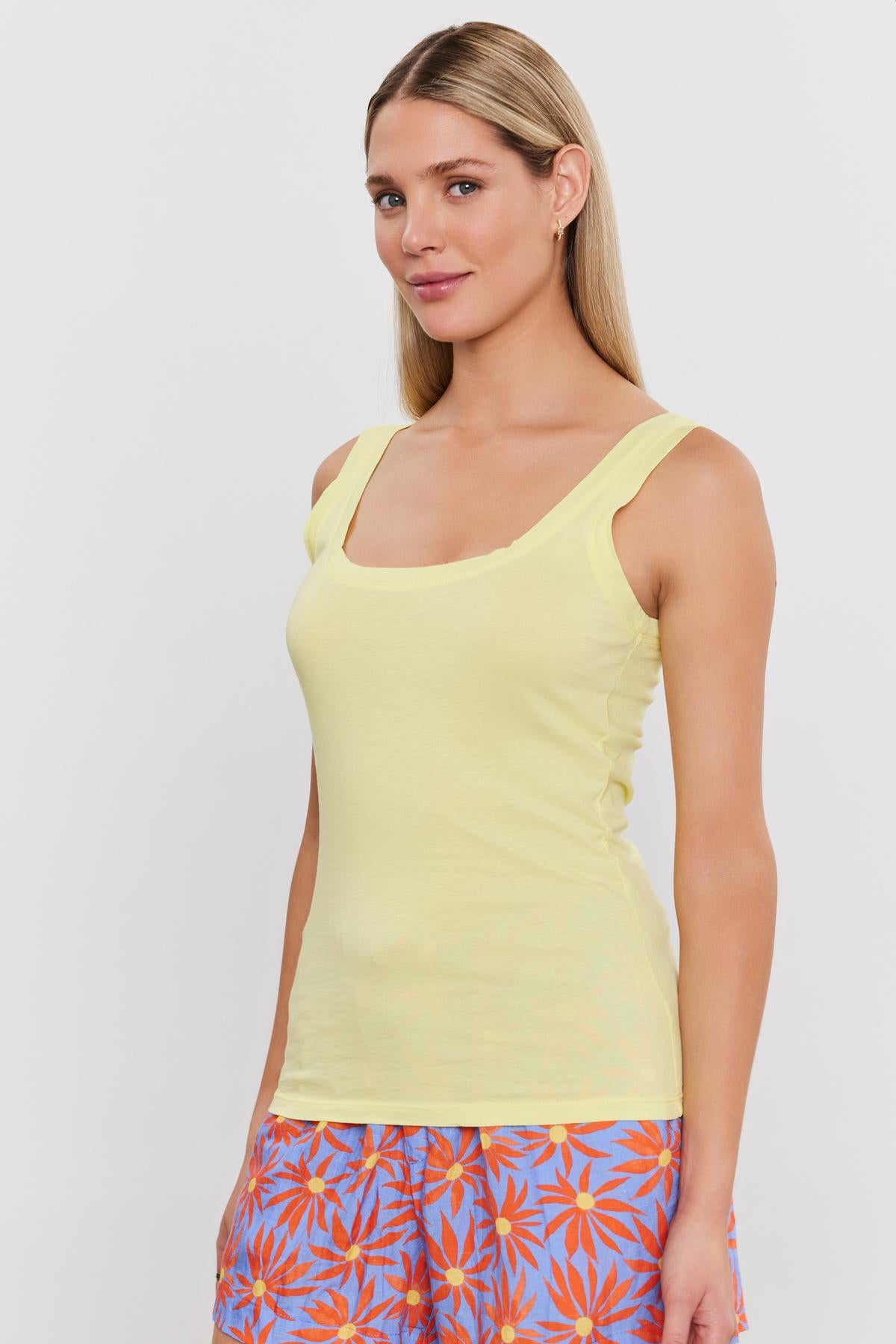 A woman wearing a yellow Velvet by Graham & Spencer MOSSY GAUZY WHISPER FITTED TANK and colorful floral shorts, standing against a white background, looking at the camera.-36910237548737