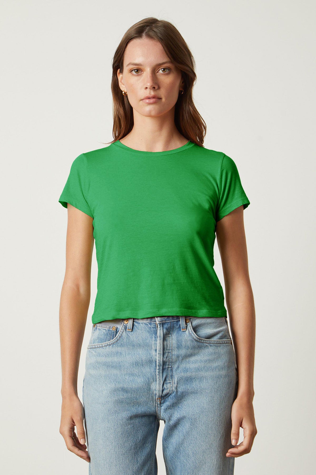   A person with long brown hair, wearing a green NINA TEE by Velvet by Graham & Spencer and blue jeans, stands against a plain background. 