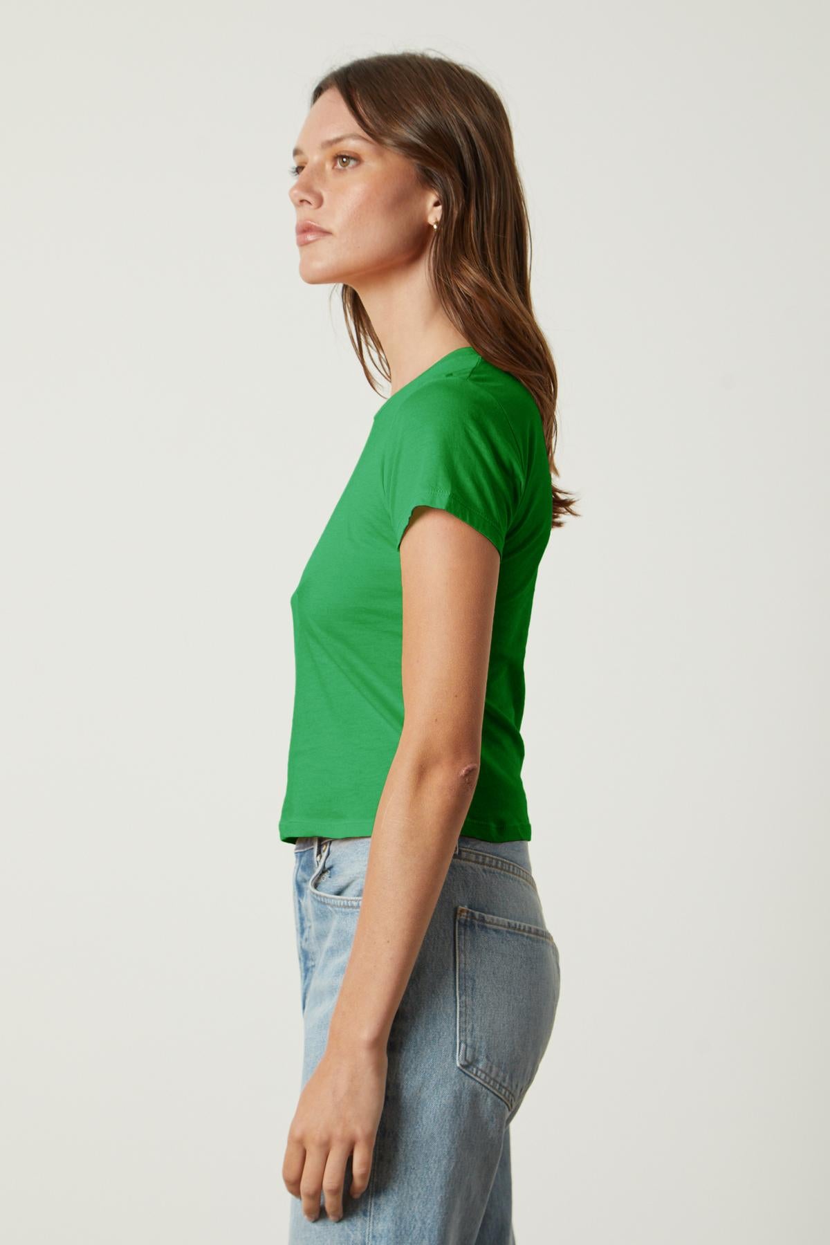   A woman stands in profile, wearing a green NINA TEE by Velvet by Graham & Spencer and blue jeans against a plain white background. 