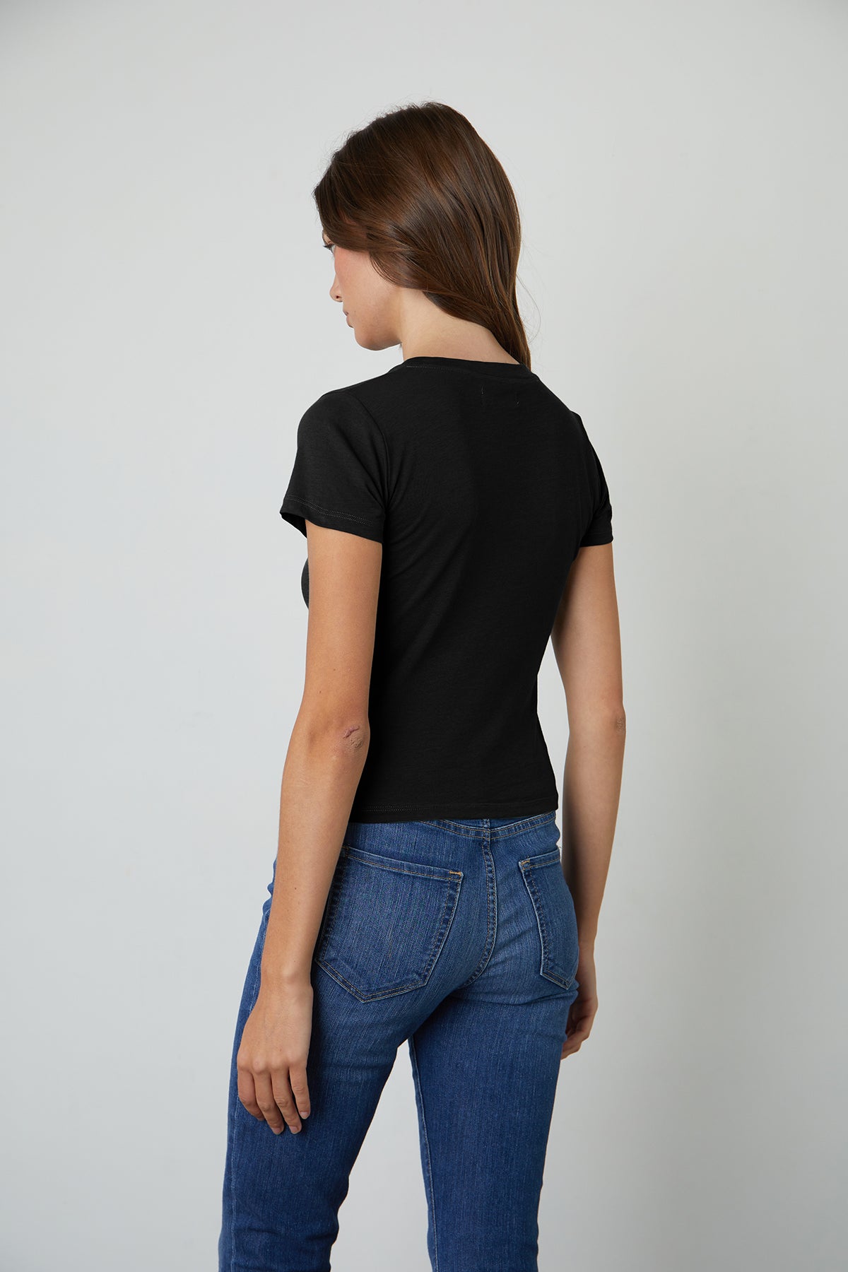   The woman is seen from behind, sporting jeans and a black t-shirt made of whisper cotton knit fabric, perfect for warm-weather, specifically the NINA CROPPED CREW NECK TEE from Velvet by Graham & Spencer. 