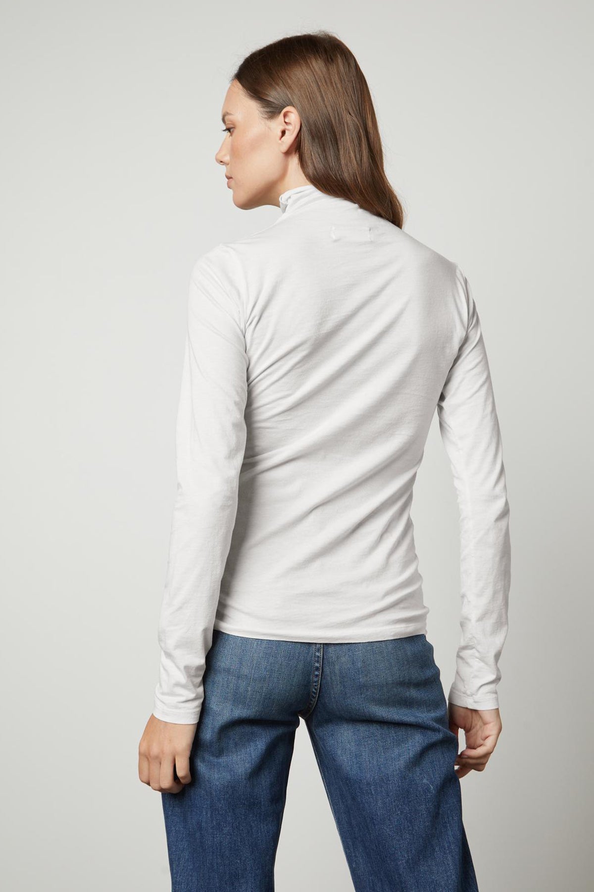   The versatile fashion world embraces the back view of a woman wearing Velvet by Graham & Spencer's TALISIA GAUZY WHISPER FITTED MOCK NECK TEE jeans and a white long-sleeved shirt. 