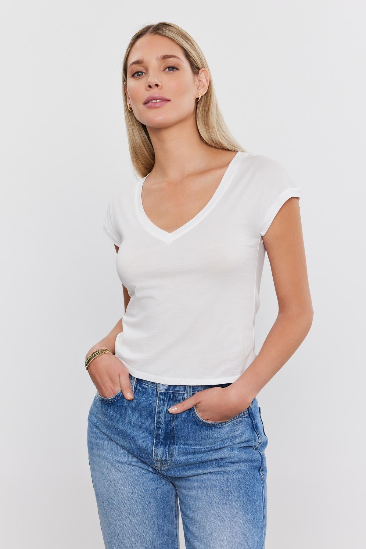 A woman wearing a white Velvet by Graham & Spencer TOBY TEE and blue jeans against a plain white background. she is posing with one hand in her pocket and looking at the camera.-36752997384385