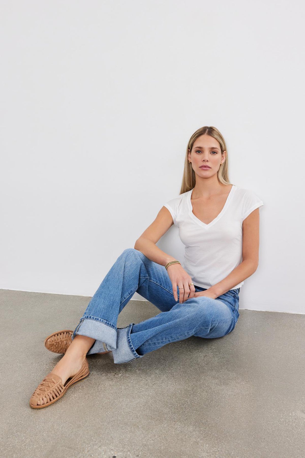   A woman in jeans and a Toby Tee by Velvet by Graham & Spencer sits on the floor against a white wall, wearing brown loafers. 