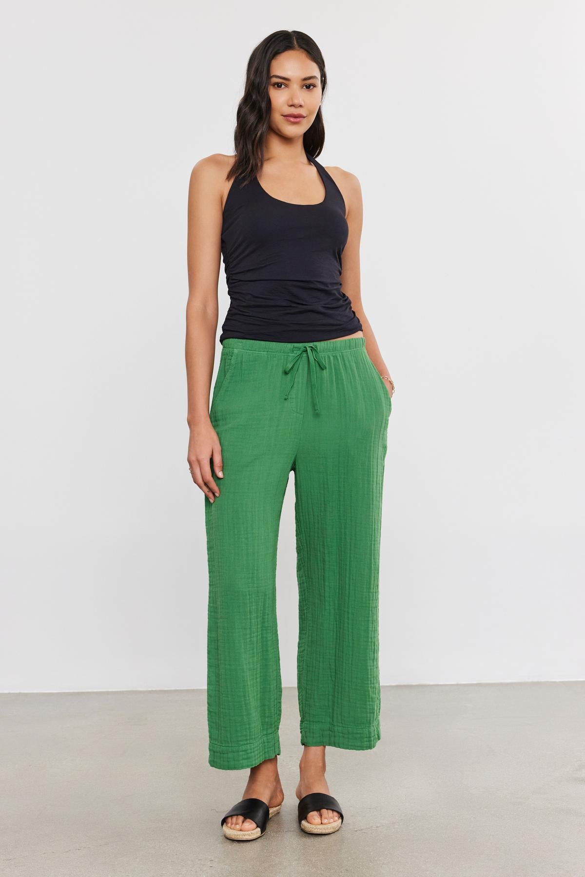 A woman stands against a plain background, wearing a black WHITLEY TANK TOP by Velvet by Graham & Spencer, green loose-fit pants, and black sandals. Her hands are in her pockets.-37402895319233