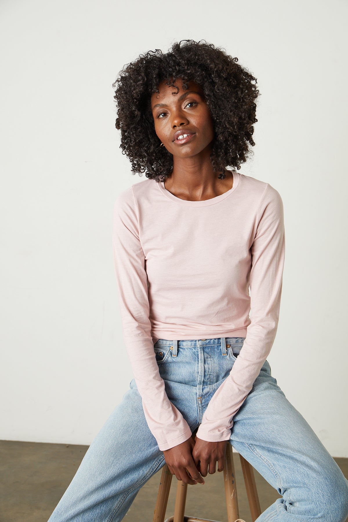 Model sitting on stool wearing Zofina Gauzy Whisper Fitted Crew Neck Tee with long sleeves in light pink ribbon color and light blue denim front-26632448835777