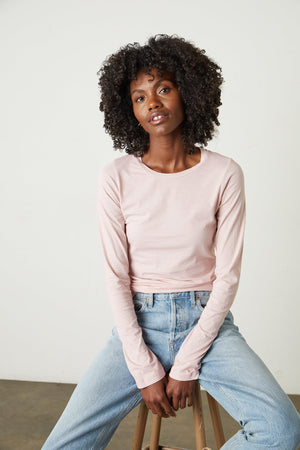 Model sitting on stool wearing Zofina Gauzy Whisper Fitted Crew Neck Tee with long sleeves in light pink ribbon color and light blue denim front