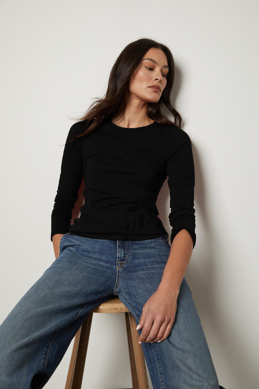 A woman sitting on a stool in black jeans and a ZOFINA GAUZY WHISPER FITTED CREW NECK TEE by Velvet by Graham & Spencer.