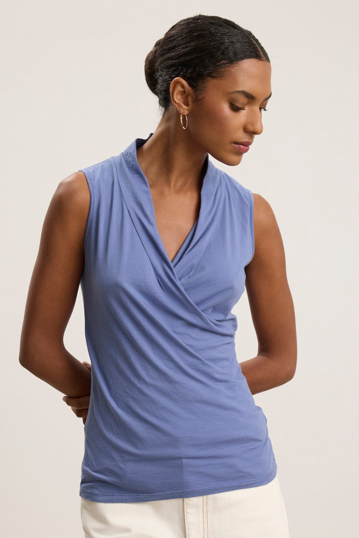 A woman models the ADELISE WRAP TANK by Velvet by Graham & Spencer, featuring a sleeveless gauzy wrap front, paired with white pants. She stands against a plain background, looking down with her hands behind her back.-39111851671745