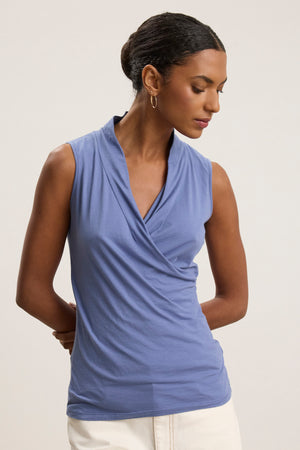 A woman models the ADELISE WRAP TANK by Velvet by Graham & Spencer, featuring a sleeveless gauzy wrap front, paired with white pants. She stands against a plain background, looking down with her hands behind her back.