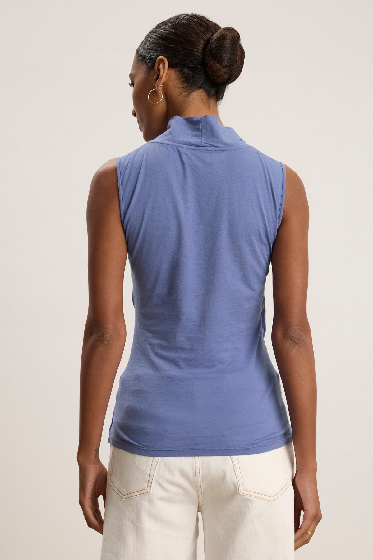A woman with her hair in a bun wears the ADELISE WRAP TANK by Velvet by Graham & Spencer in light blue, paired with white jeans, shown from the back against a plain background.-39111851638977