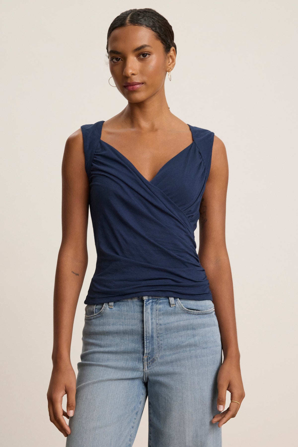 A woman stands against a plain light background wearing the Velvet by Graham & Spencer AZURA TANK TOP, a sleeveless navy blue wrap with a romantic front crossover, paired with light blue jeans.-39119703670977